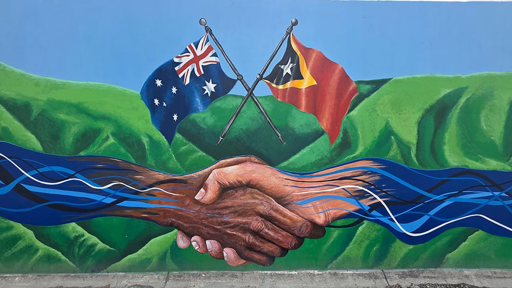 mural of australian flag and timorese flag above two hands shaking. the green of mountains in the background